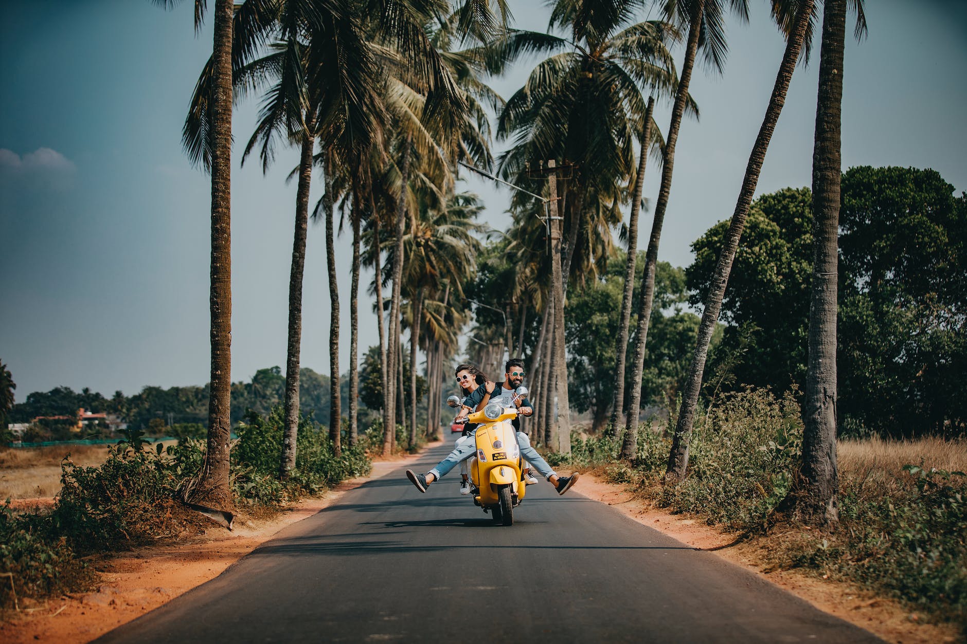 A boy and girl ride a motorbike alongside a beach with palm trees 